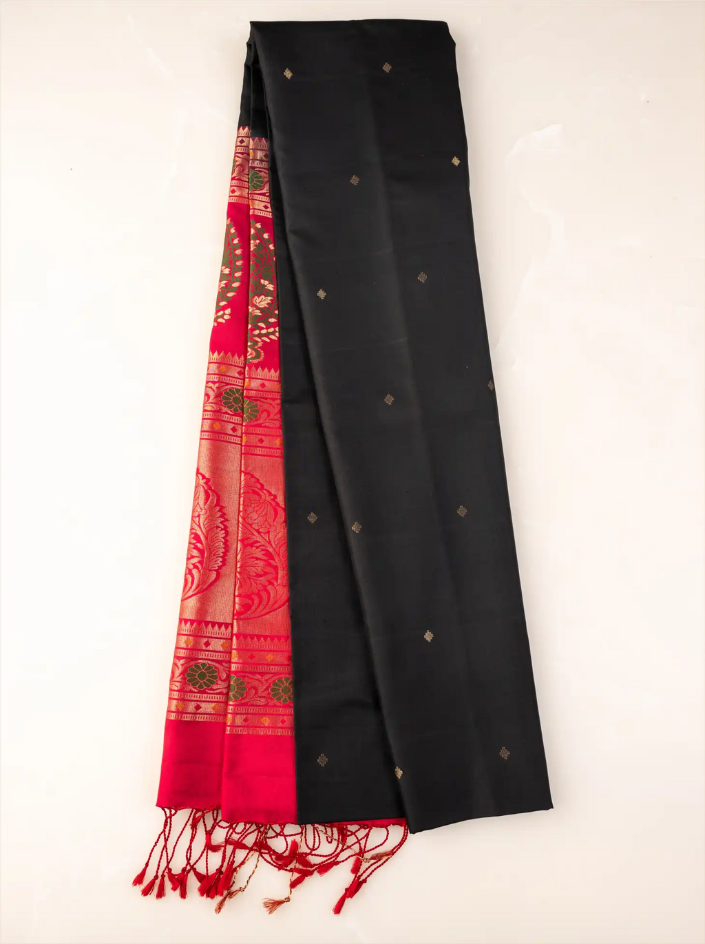 Neatly folded handwoven jet black pure Paithani silk saree showcasing small gold zari buttis, a vibrant red pallu with intricate peacock weave motifs, and red tassels along the border.
