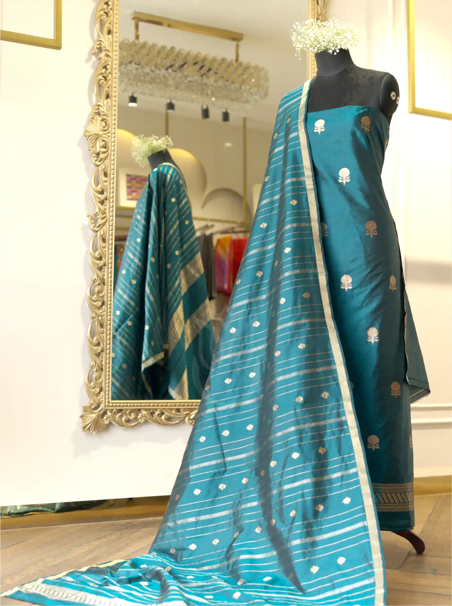 Teal blue handwoven Chinia silk suit set displayed on a mannequin, featuring gold zari floral bootis, a striped zari tissue dupatta with booti details, and a large ornate gold mirror in the background.