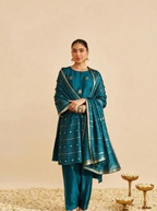 Woman posing in a handwoven teal blue Chinia silk suit set featuring woven gold zari floral bootis and a striped zari dupatta against a beige background.