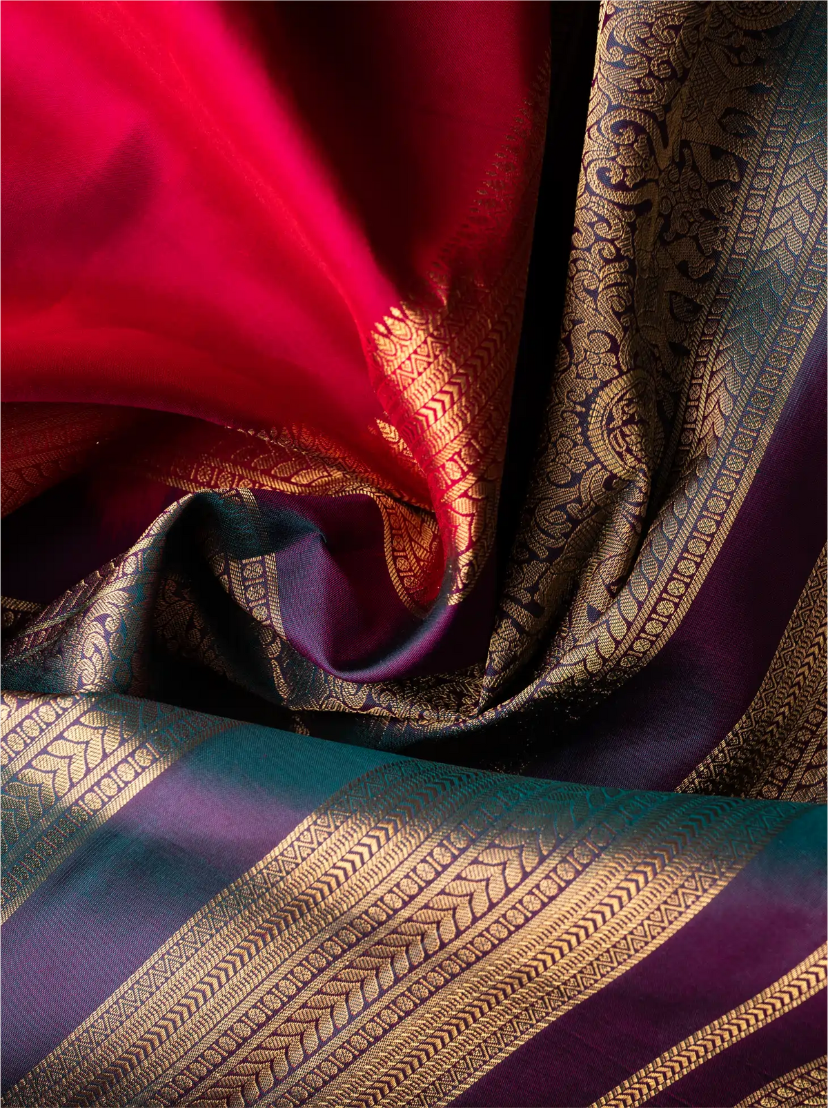 Close-up view of a handwoven crimson red Kanjivaram silk saree draped in soft folds, featuring fine gold zari motifs paired with an intricate deep teal border and pallu woven with rich geometric and floral zari patterns.