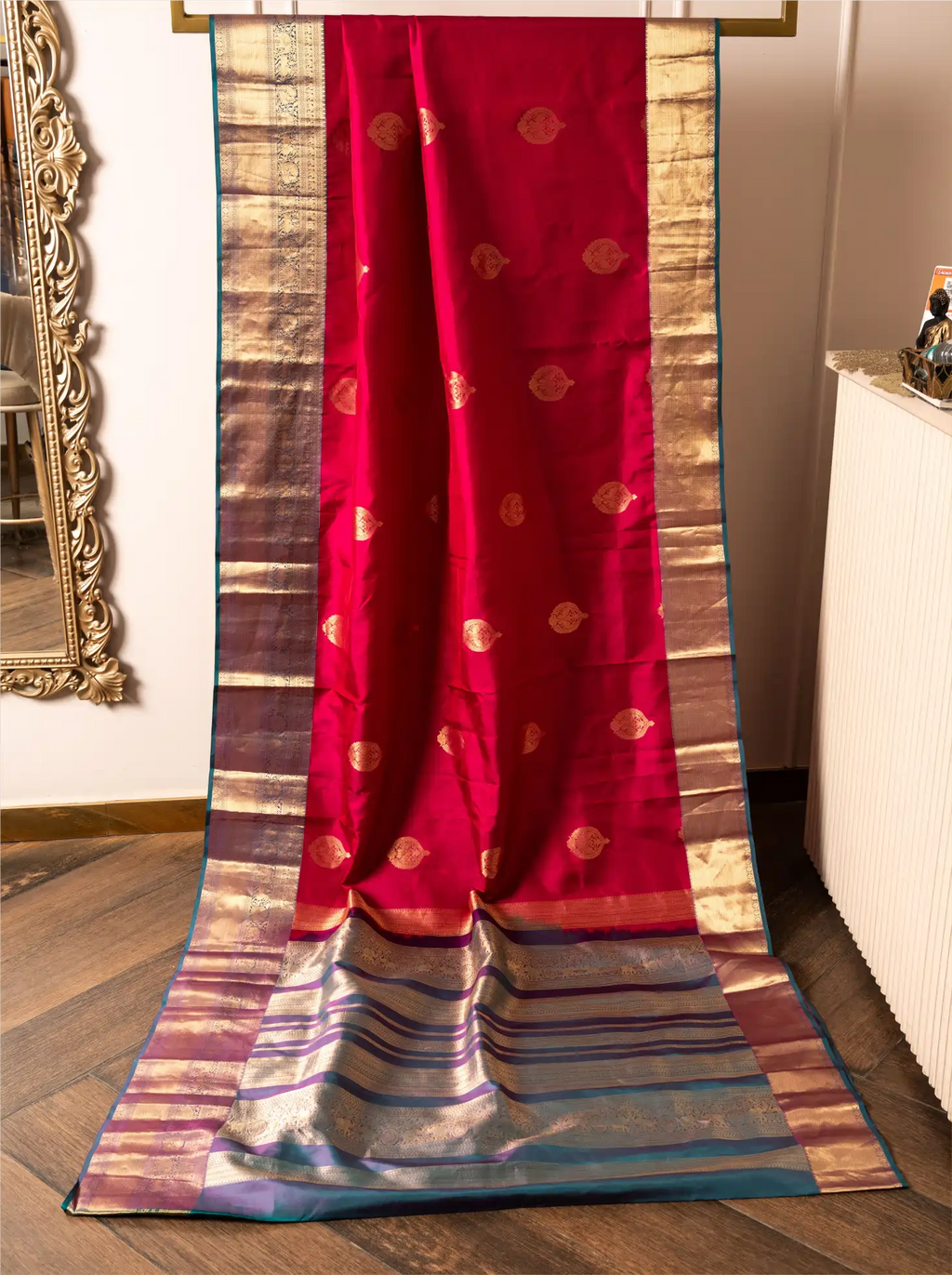 Handwoven crimson red pure Kanjivaram silk saree displayed vertically from a rack, featuring gold zari motifs on the body, broad gold and teal selvedge borders, and a decorative contrast teal pallu flowing to the floor.