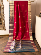 Handwoven crimson red pure Kanjivaram silk saree displayed vertically from a rack, featuring gold zari motifs on the body, broad gold and teal selvedge borders, and a decorative contrast teal pallu flowing to the floor.