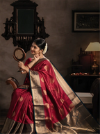 Model seated in a traditional interior setting wearing a handwoven crimson red pure Kanjivaram silk saree adorned with gold zari motifs, draped with a rich contrast teal border and intricate brocade pallu.