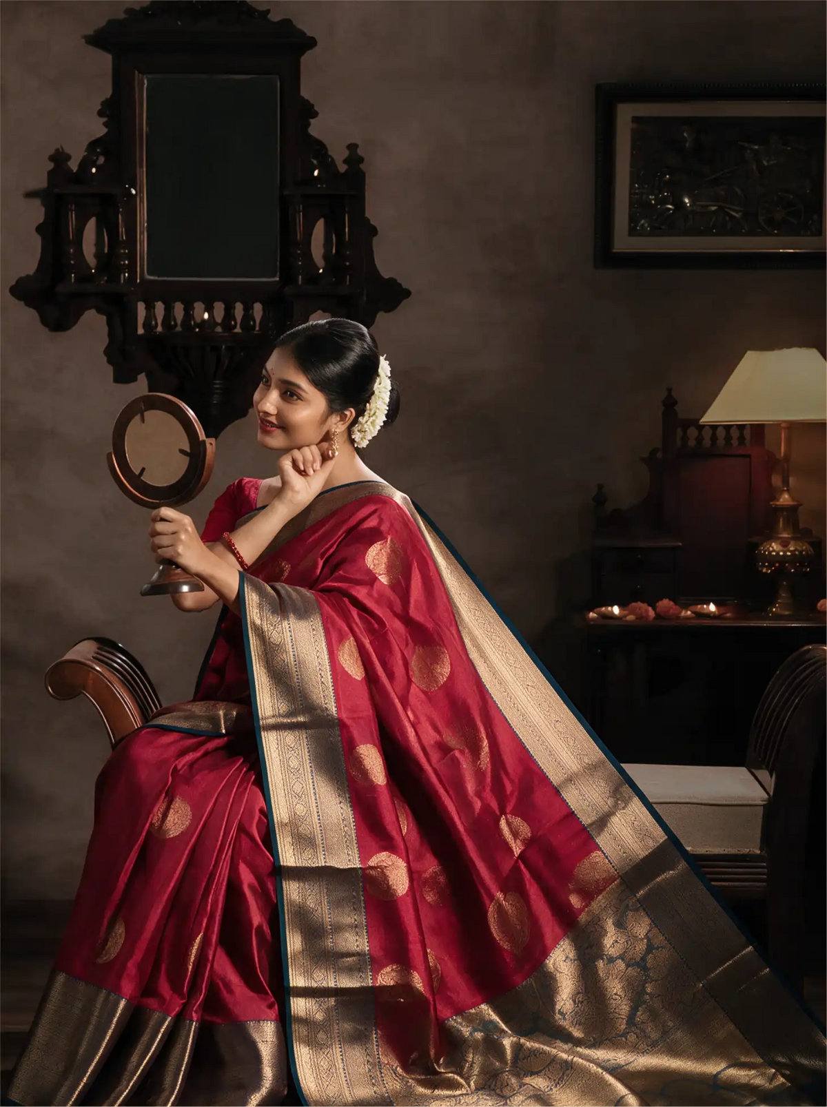Model seated in a traditional interior setting wearing a handwoven crimson red pure Kanjivaram silk saree adorned with gold zari motifs, draped with a rich contrast teal border and intricate brocade pallu.