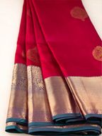 Close-up view of a handwoven crimson red pure Kanjivaram silk saree showcasing neatly draped pleats, traditional gold zari floral motifs on the body, and a contrasting deep teal border woven with fine gold brocade.
