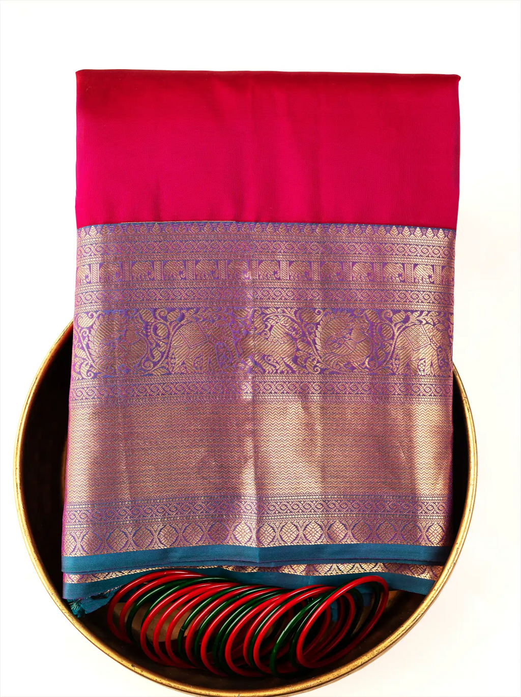 Handwoven crimson red pure Kanjivaram silk saree folded inside a decorative brass tray with matching green and red bangles, showcasing its gold zari woven pallu and deep teal border.