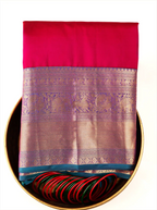 Handwoven crimson red pure Kanjivaram silk saree folded inside a decorative brass tray with matching green and red bangles, showcasing its gold zari woven pallu and deep teal border.