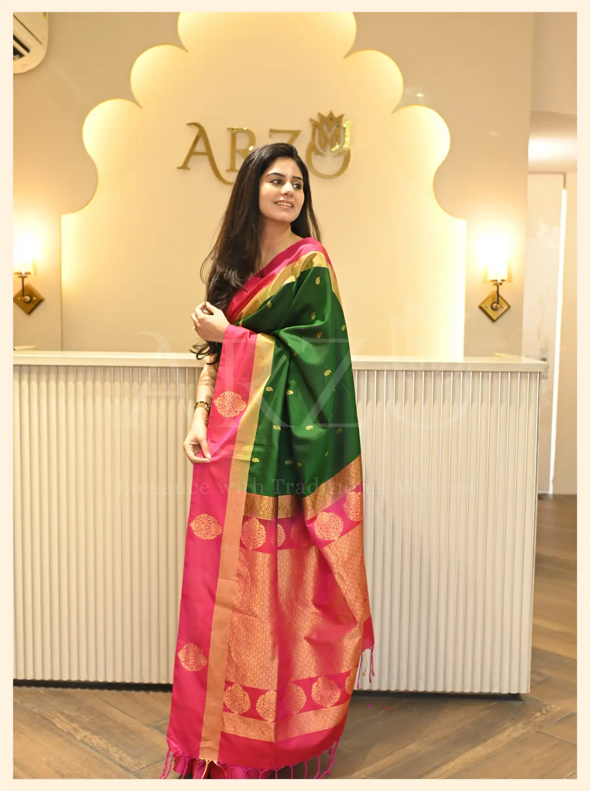 Woman posing in a handwoven dark green Mysore silk saree with a pink border and gold zari pattern from Arzo.