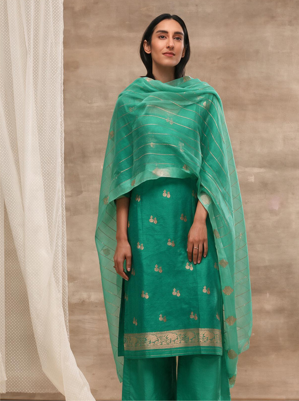 Woman modeling an emerald green handwoven silk suit set with gold zari floral bootis, styled with a draped sheer striped matching dupatta against a neutral backdrop