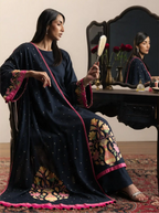Woman sitting in a dressing room holding a hand mirror, wearing a handwoven pure georgette silk charcoal black suit set with micro bootis, contrast pink piping, and a multi-color floral woven dupatta.