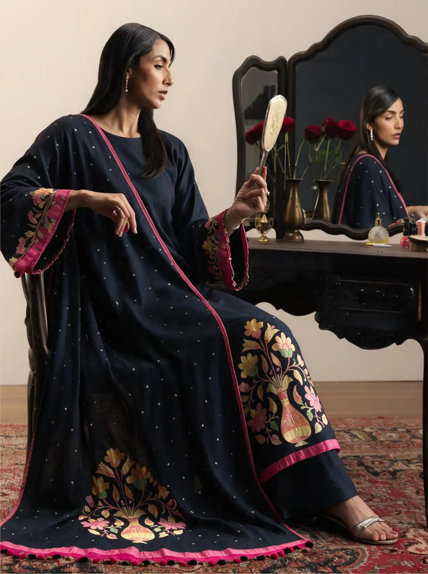 Woman sitting in a dressing room holding a hand mirror, wearing a handwoven pure georgette silk charcoal black suit set with micro bootis, contrast pink piping, and a multi-color floral woven dupatta.