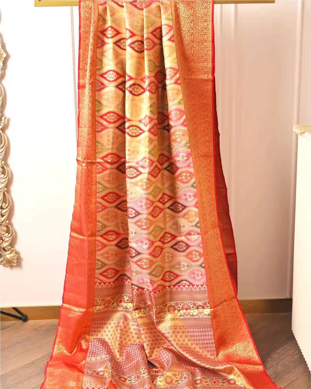 Handwoven champagne gold and coral orange Rangkart Kanchipuram silk saree draped on a display stand showing geometric zari motifs and rich border.