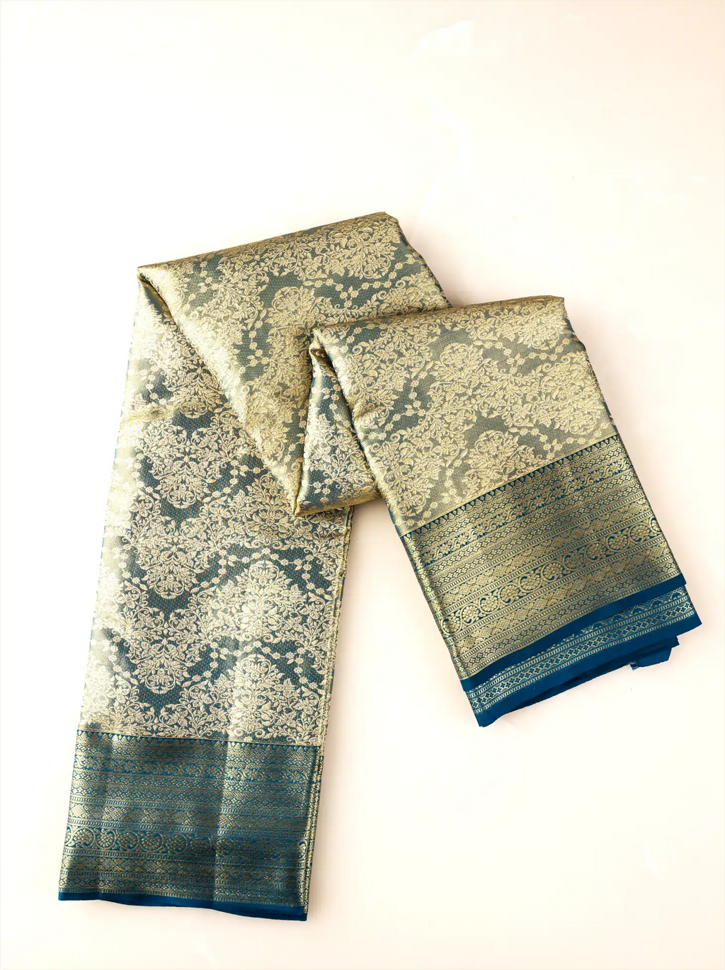 Handwoven metallic gold tissue Kanjivaram silk saree elegantly folded and draped to showcase its intricate dark teal zari brocade patterns across the main body, paired with a solid dark teal border with woven gold zari highlights.