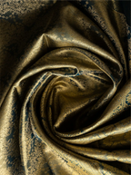 Macro shot of a handwoven tissue Kanjivaram silk saree arranged in a soft circular swirl, highlighting the rich metallic gold shimmer of the fabric alongside intricate dark teal zari brocade floral weave patterns.