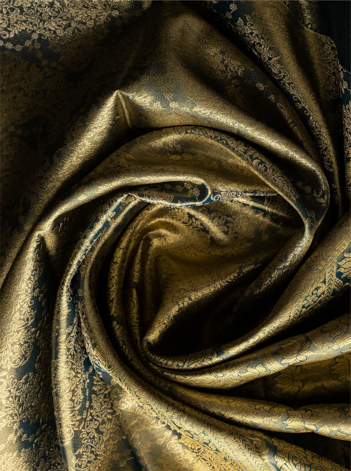 Macro shot of a handwoven tissue Kanjivaram silk saree arranged in a soft circular swirl, highlighting the rich metallic gold shimmer of the fabric alongside intricate dark teal zari brocade floral weave patterns.