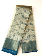 Handwoven metallic gold tissue Kanjivaram silk saree folded neatly against a white background, displaying intricate dark teal zari brocade floral patterns across the body and a solid dark teal border.