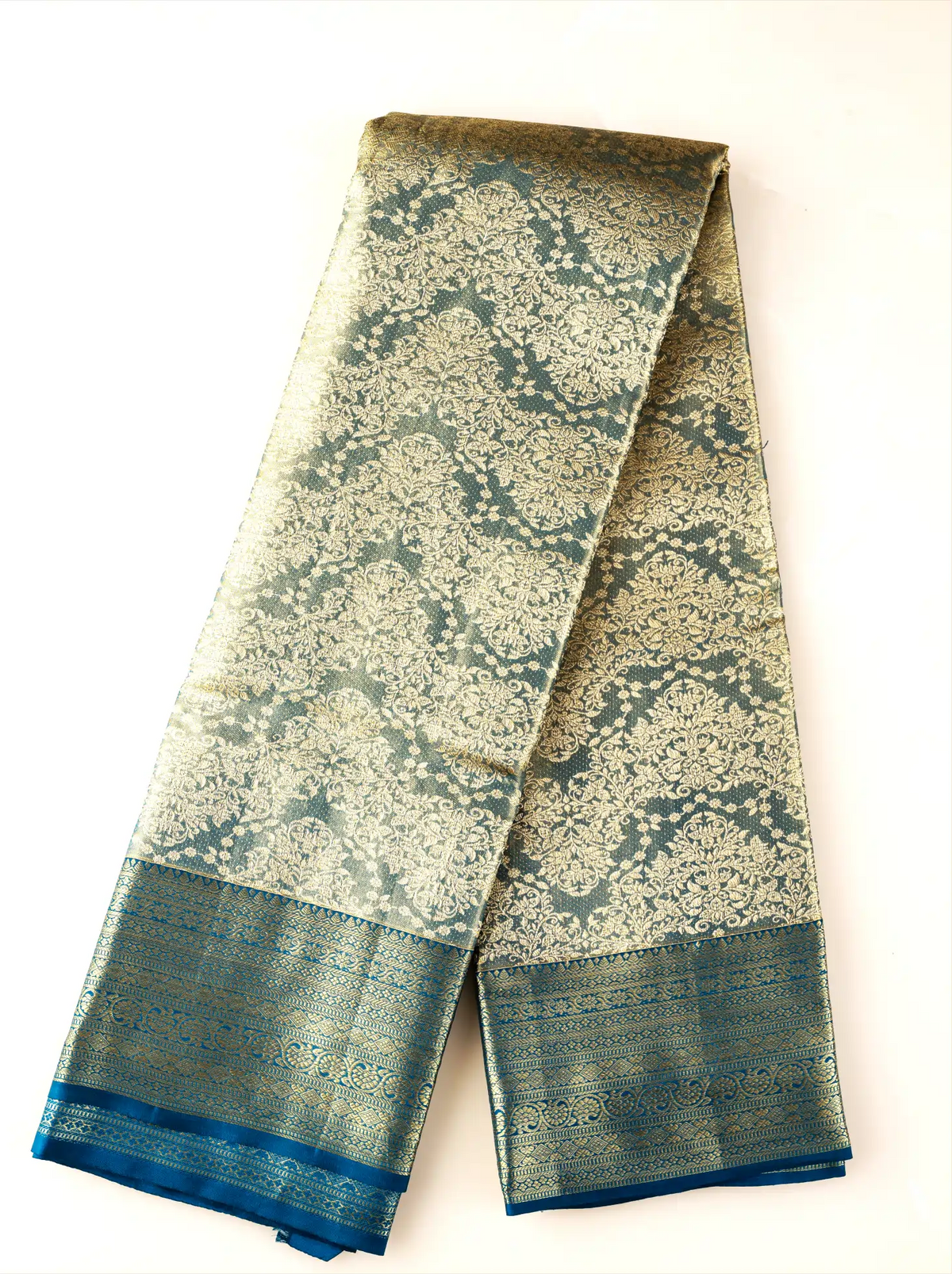 Handwoven metallic gold tissue Kanjivaram silk saree folded neatly against a white background, displaying intricate dark teal zari brocade floral patterns across the body and a solid dark teal border.
