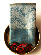Handwoven metallic gold tissue Kanjivaram silk saree folded neatly on a brass tray, featuring ornate teal zari brocade weave patterns paired with red and green glass bangles.