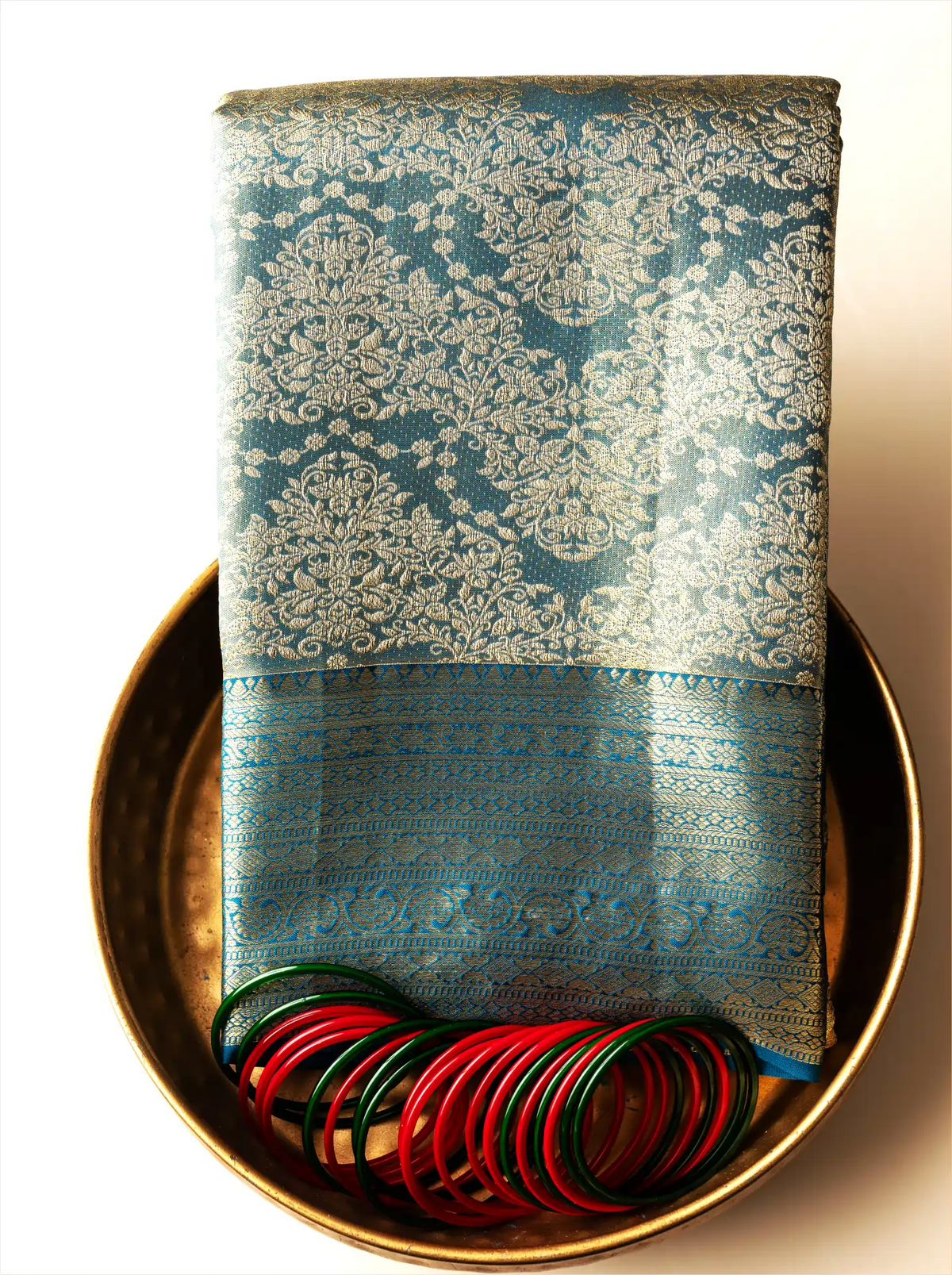 Handwoven metallic gold tissue Kanjivaram silk saree folded neatly on a brass tray, featuring ornate teal zari brocade weave patterns paired with red and green glass bangles.
