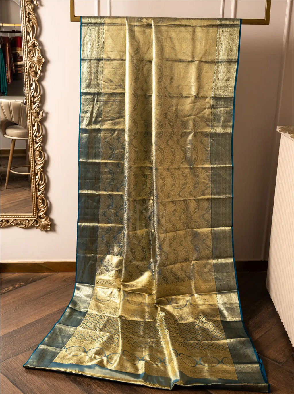 Handwoven metallic gold tissue Kanjivaram silk saree displayed vertically from a rack, showcasing its full pleated drape, rich metallic shimmer, dark teal zari border, and decorative lower pallu weave.