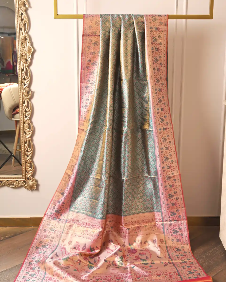 Handwoven green tissue Patola Kanjivaram silk saree with pink floral zari border draped on a gold display rack next to a decorative mirror.