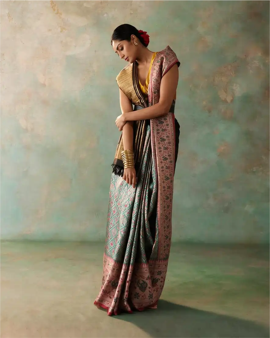 Woman wearing a handwoven green tissue Patola Kanjivaram silk saree with a pink zari floral border and gold blouse, posing in a studio.