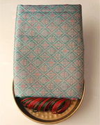 Folded sea green tissue Patola Kanjivaram silk saree with intricate geometric lattice weave presented on a brass tray alongside red and green glass bangles.