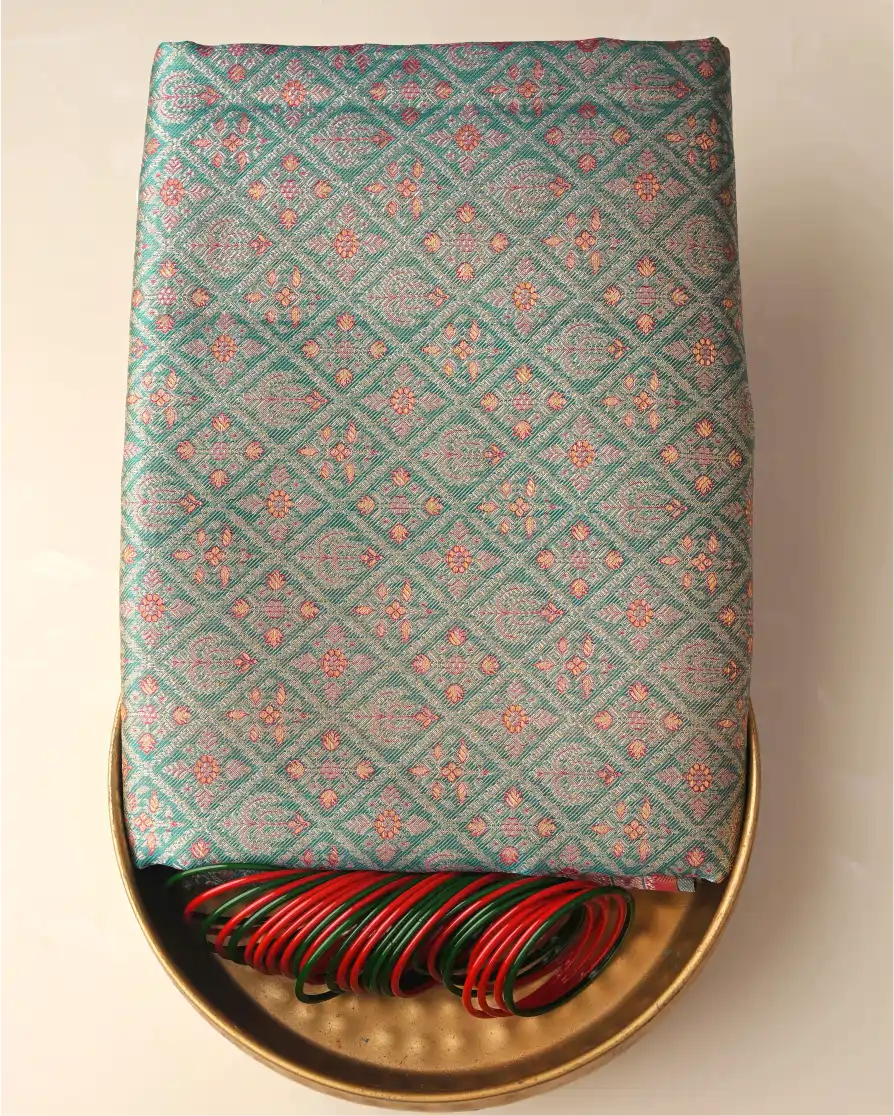 Folded sea green tissue Patola Kanjivaram silk saree with intricate geometric lattice weave presented on a brass tray alongside red and green glass bangles.