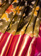 Close-up view of a folded handwoven Kanjivaram Patola silk saree showing a black, olive green, and yellow ikat patterned body alongside a vibrant metallic magenta-pink zari border.