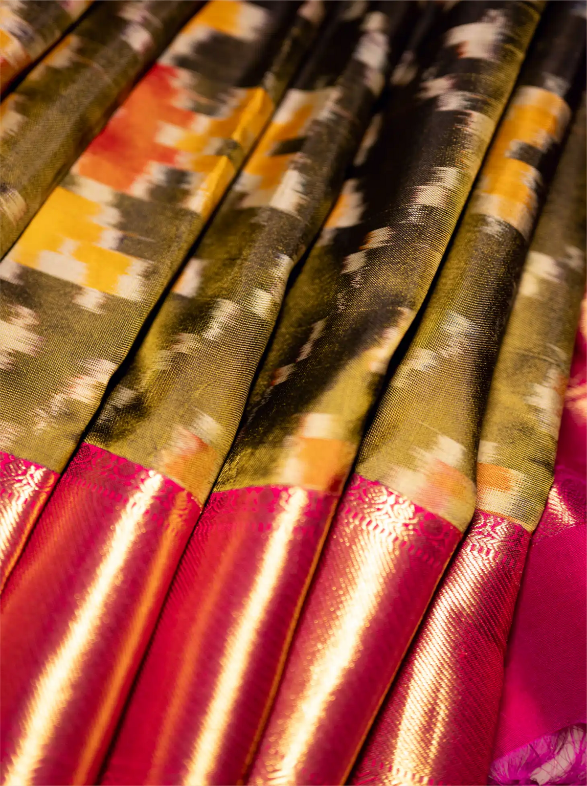 Close-up view of a folded handwoven Kanjivaram Patola silk saree showing a black, olive green, and yellow ikat patterned body alongside a vibrant metallic magenta-pink zari border.