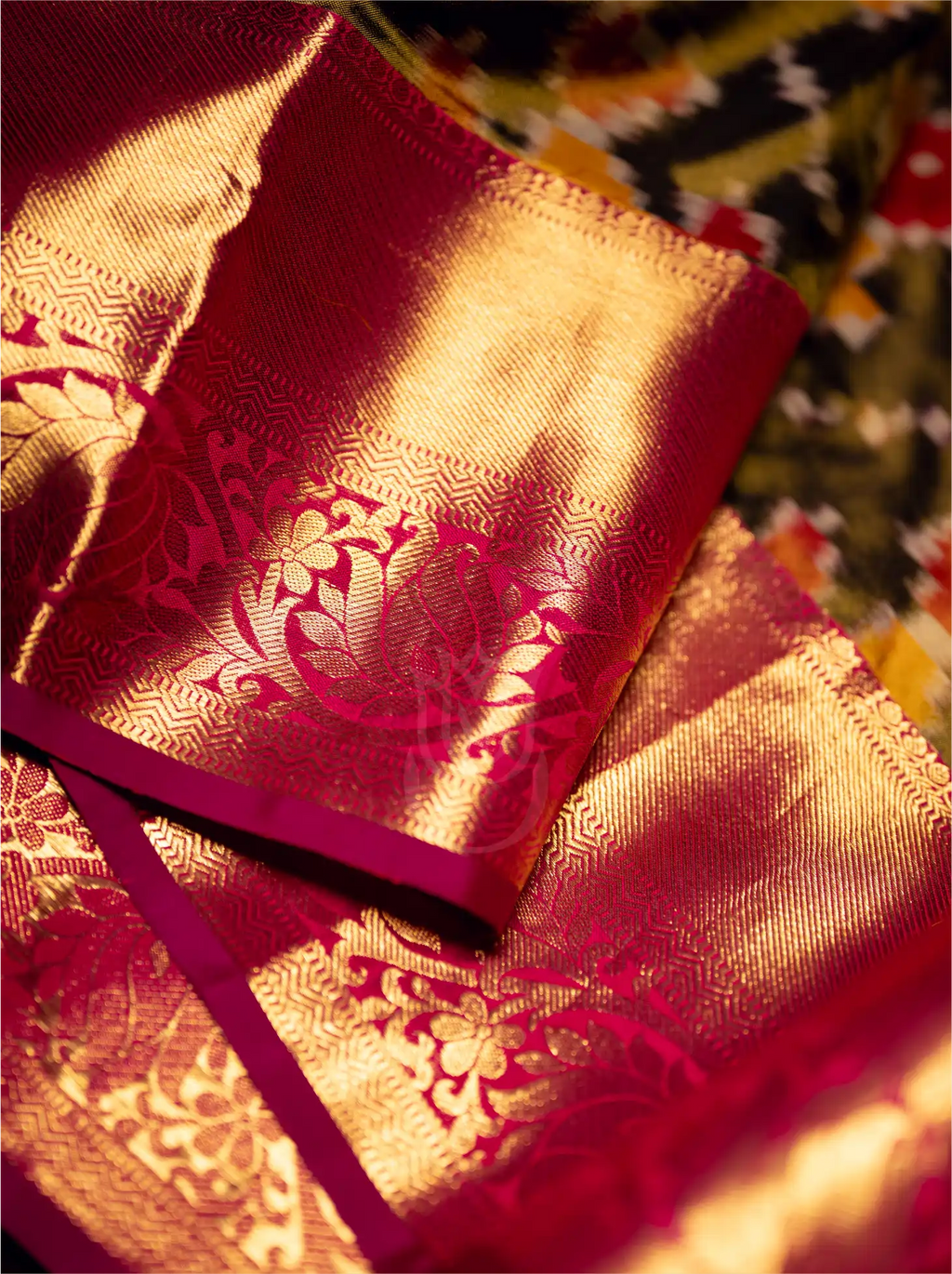 Close-up shot of a folded handwoven Kanjivaram Patola silk saree showcasing a rich crimson red pallu with elaborate gold zari floral motifs and a detailed woven border.