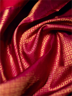 Close-up view of the folded crimson red handwoven Kanjivaram Patola silk saree showcasing rich metallic gold zari weaving with intricate square-grid brocade patterns.