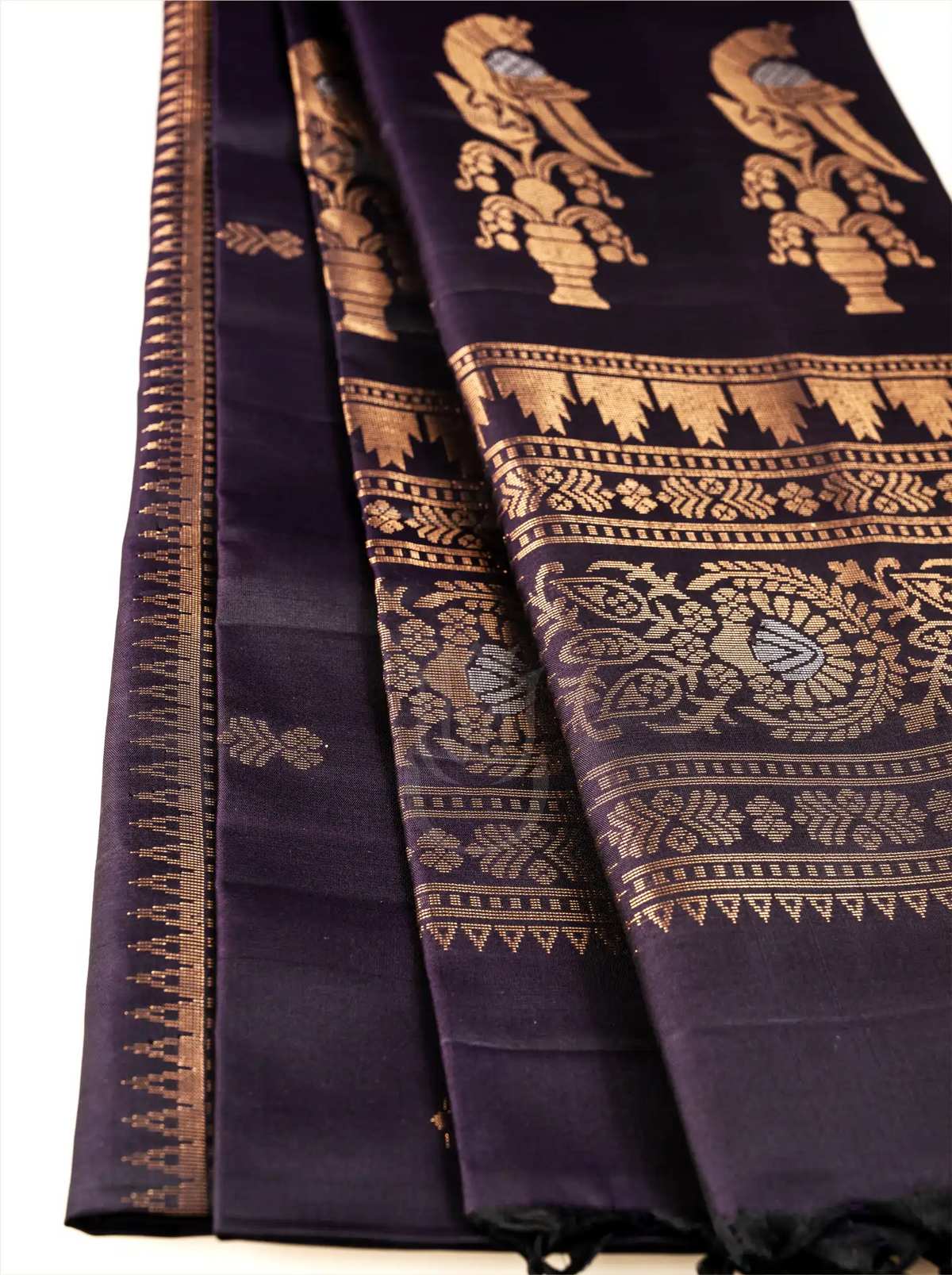 Close-up shot of a deep royal aubergine Kanjivaram silk saree showing delicate gold zari bootis on the pleats, paired with an intricately woven zari pallu featuring gold bird and floral motifs on a dark backdrop
