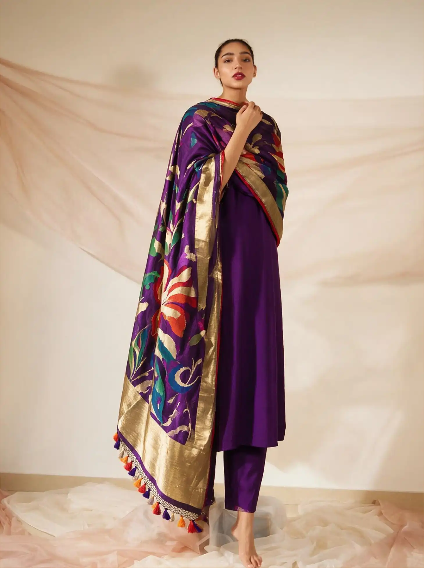 Full-length view of a model wearing a royal purple handwoven Katan silk suit, styled with a draped purple dupatta featuring multi-colored woven floral motifs, broad gold zari borders, and tassel fringe against a draped beige backdrop