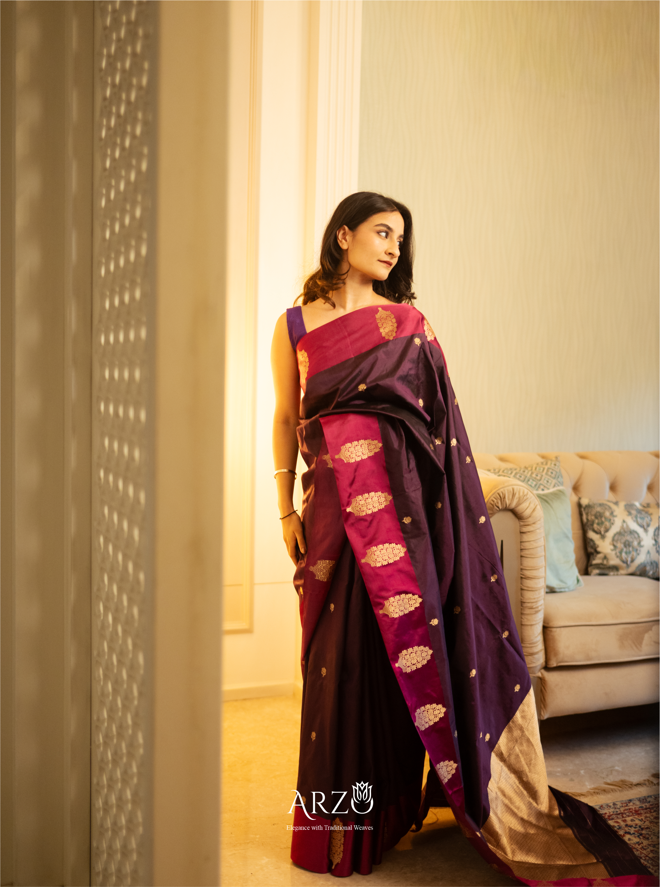 Woman wearing a purple saree with a pattern, standing in a room with a sofa and decorative pillows.