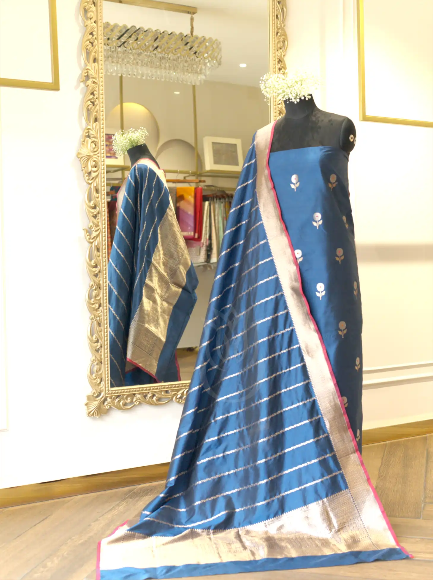 Royal blue handwoven Kora Katan silk suit set displayed on a mannequin, featuring silver booti work, a striped gold zari tissue dupatta with a pink border, and a large ornate gold mirror in the background.