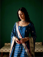 Woman posing in a handwoven royal blue Kora Katan silk suit set with silver floral bootis and a gold zari striped dupatta featuring a contrast pink border against a green backdrop.