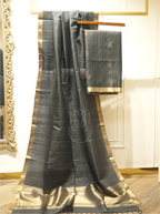 Charcoal black handwoven Matka silk dupatta with gold zari borders draped alongside folded suit fabric on a wooden hanger, set against an ornate gold mirror