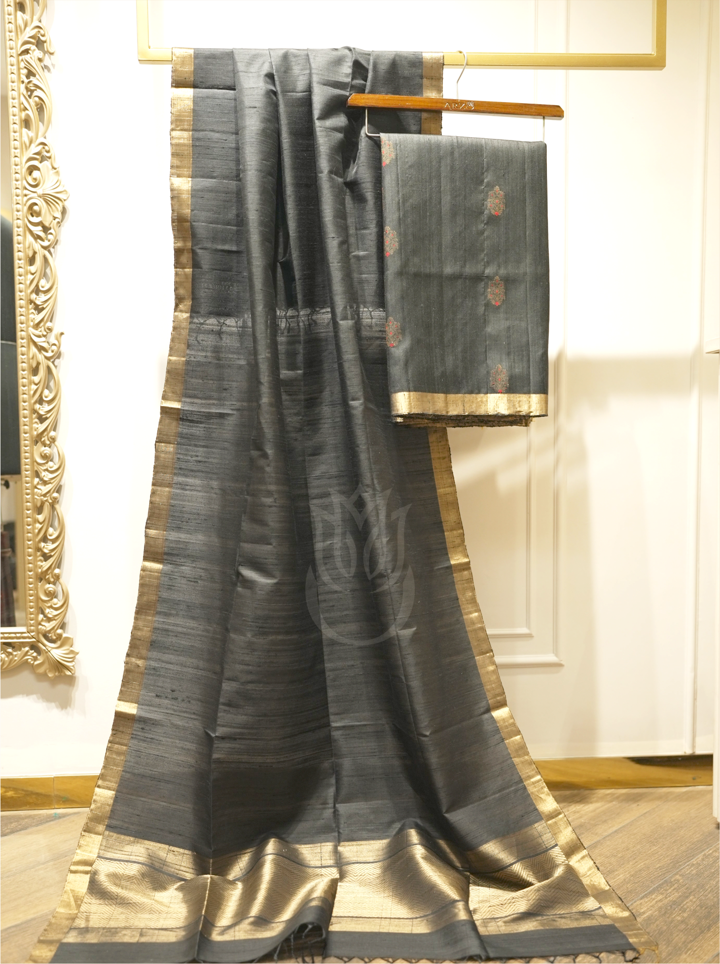 Charcoal black handwoven Matka silk dupatta with gold zari borders draped alongside folded suit fabric on a wooden hanger, set against an ornate gold mirror
