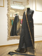 Full-length display of a charcoal black handwoven Matka silk suit on a mannequin, showcasing a draped dupatta with an antique gold zari border reflected in a decorative mirror