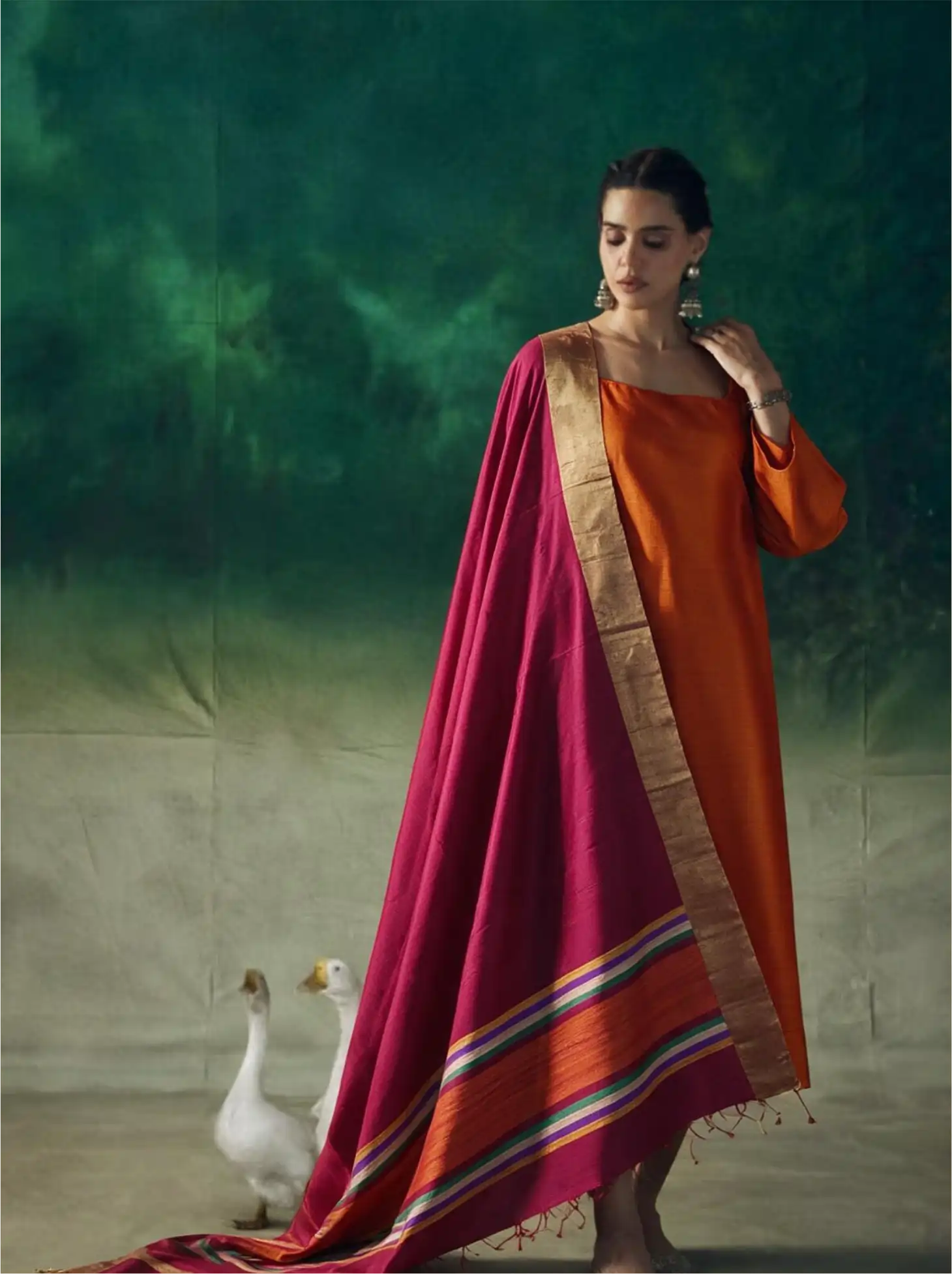Woman modeling a vibrant orange raw silk suit paired with a deep magenta Matka silk dupatta featuring gold borders and multi-color striped palla