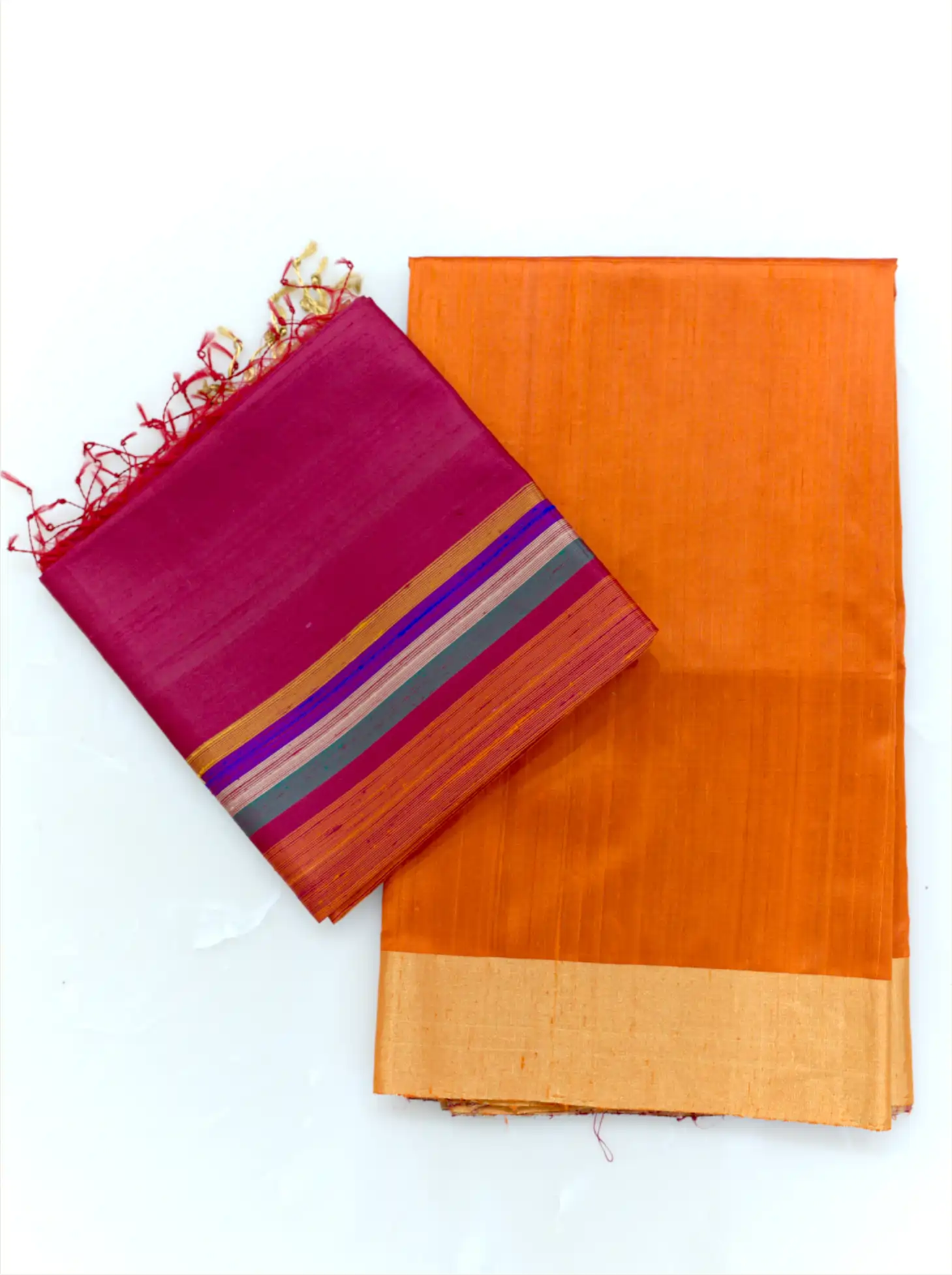 Neatly folded orange raw silk fabric featuring a gold tissue border paired with a folded magenta dupatta displaying vibrant multi-color stripes and fringe tassels on a plain background