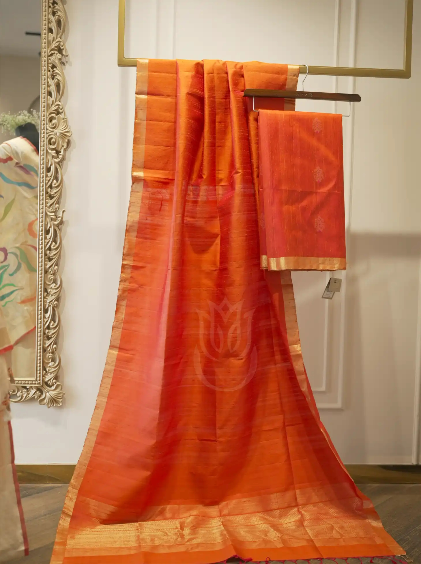 Rust orange handwoven Matka silk dupatta with wide gold zari tissue borders, displayed vertically alongside folded suit fabric on a wooden hanger near an ornate mirror