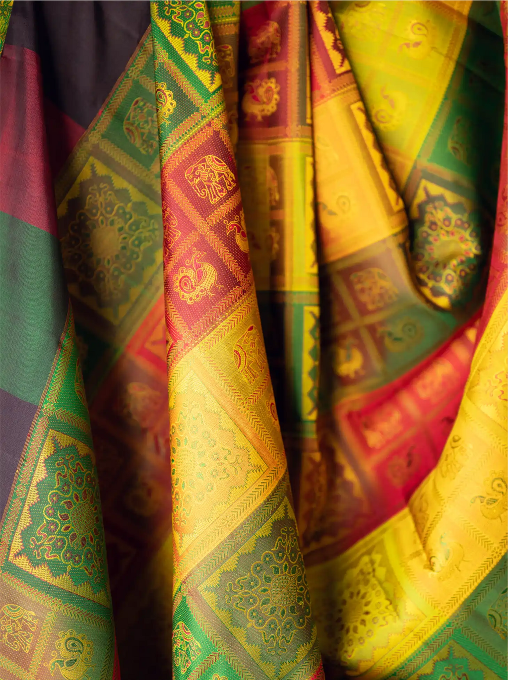 Close-up view of a handwoven brocade silk saree displaying rich golden-yellow, emerald green, crimson red, and purple draped folds with metallic zari accents and intricate traditional woven motifs.