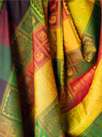 Close-up view of a handwoven brocade silk saree displaying rich golden-yellow, emerald green, crimson red, and purple draped folds with metallic zari accents and intricate traditional woven motifs.
