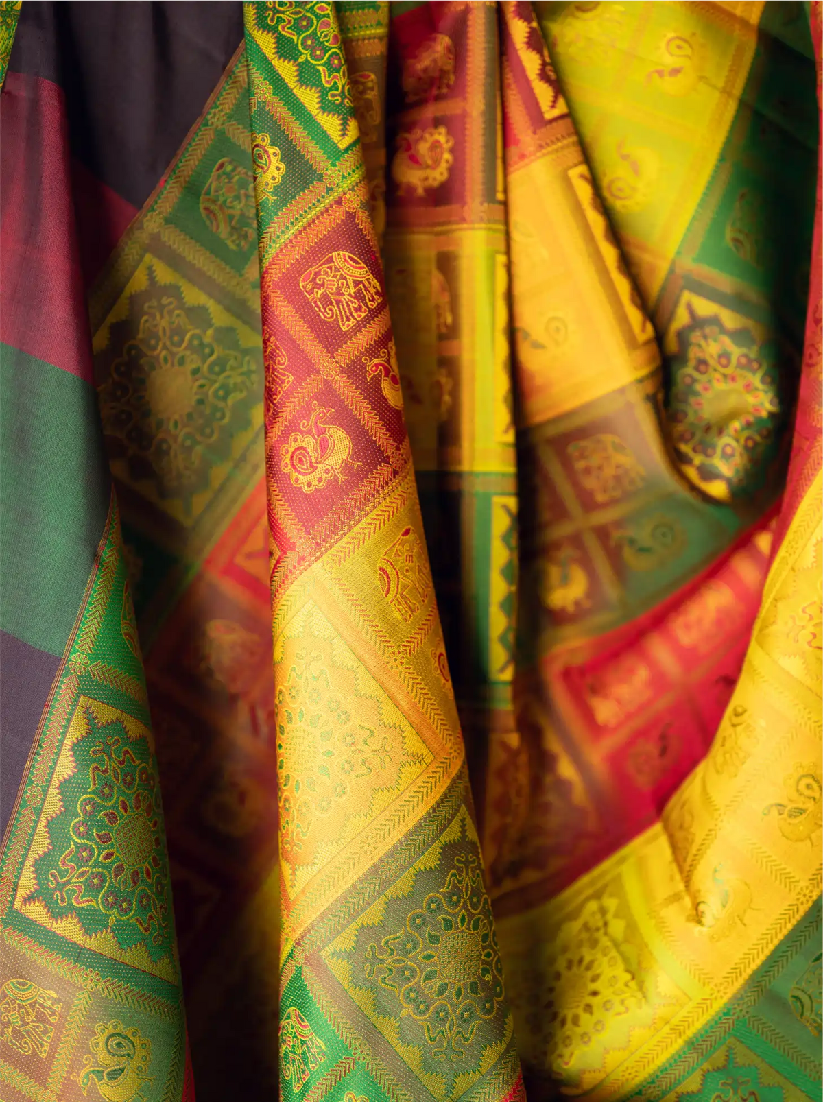 Close-up view of a handwoven brocade silk saree displaying rich golden-yellow, emerald green, crimson red, and purple draped folds with metallic zari accents and intricate traditional woven motifs.