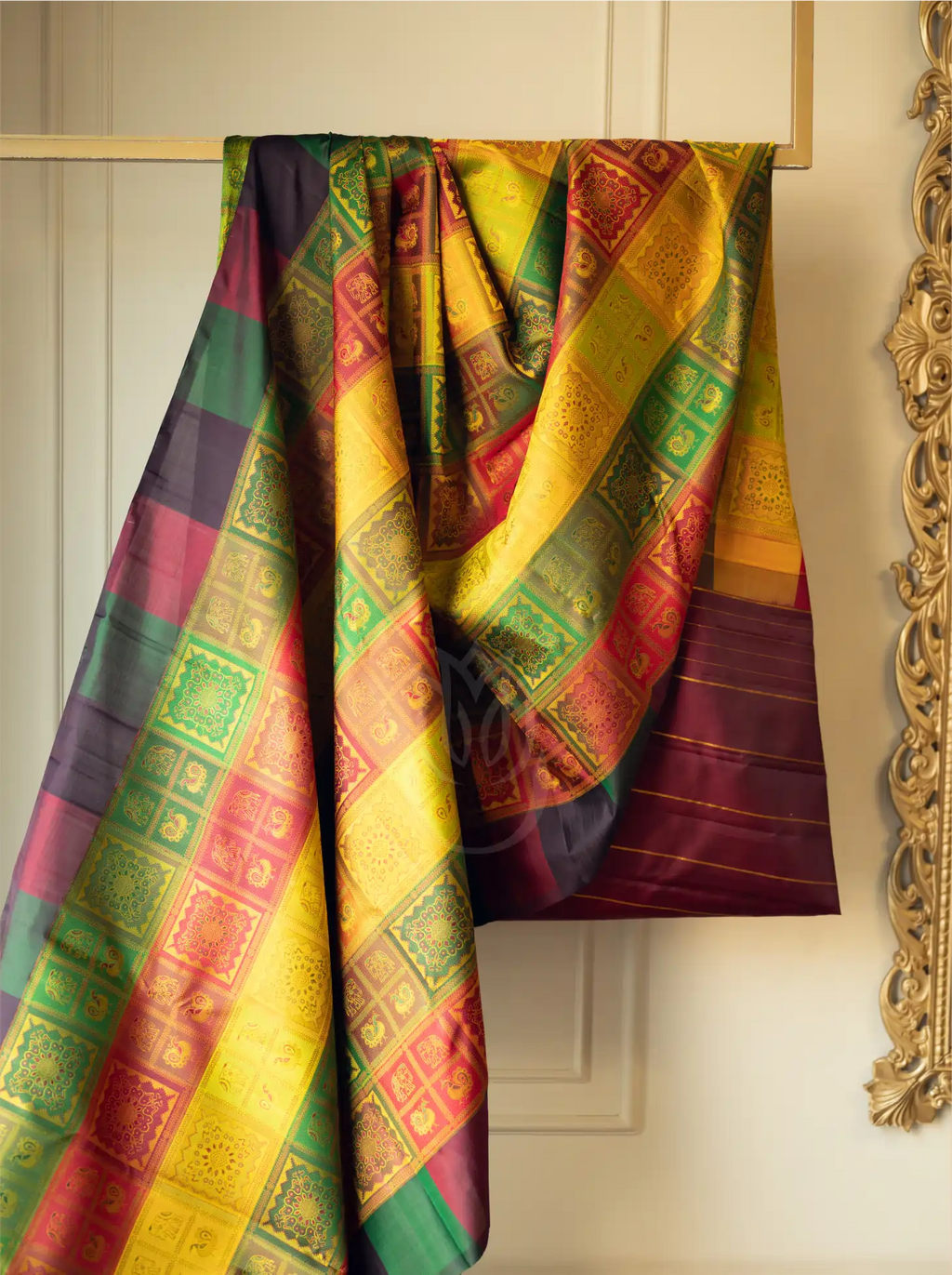 A handwoven multicolor brocade silk saree draped over a wooden hanger, featuring intricate golden zari block patterns across vibrant panels of yellow, green, red, and purple against a cream wall.