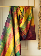 A handwoven multicolor brocade silk saree draped over a wooden hanger, featuring intricate golden zari block patterns across vibrant panels of yellow, green, red, and purple against a cream wall.