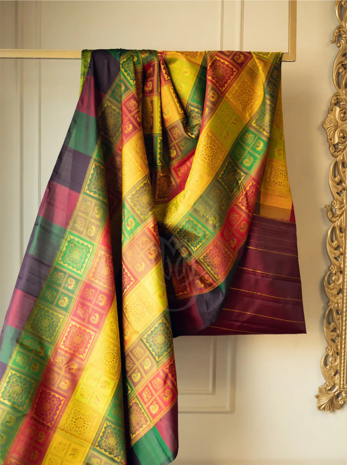 A handwoven multicolor brocade silk saree draped over a wooden hanger, featuring intricate golden zari block patterns across vibrant panels of yellow, green, red, and purple against a cream wall.