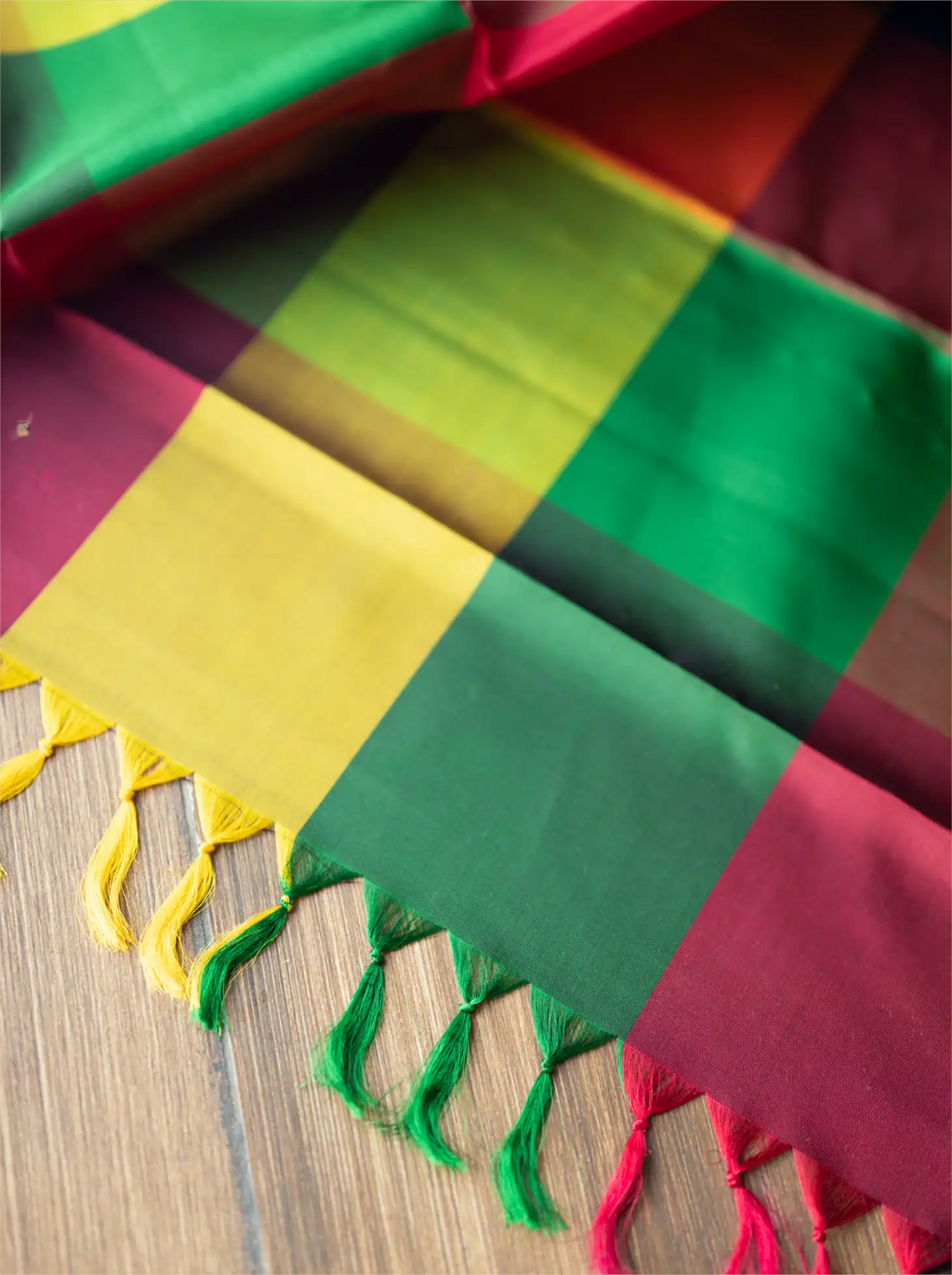 Close-up detail of a handwoven multicolor brocade silk saree pallu showcasing vibrant green, mustard yellow, and magenta red woven blocks with handcrafted yellow and green fringe tassels along the edge on a wooden surface.